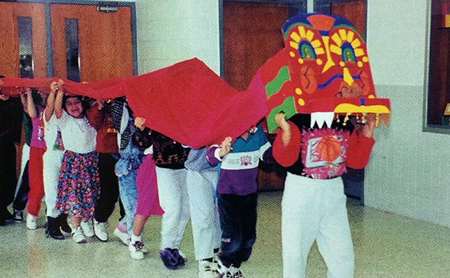 Color photograph from our 1993 to 1994 yearbook showing students participating in a Lion or Dragon Dance. The students are lined up in a row, underneath a long tail of red fabric extending from an intricately painted lion or dragon head worn by the student at the front of the group. The children are parading through a hallway.