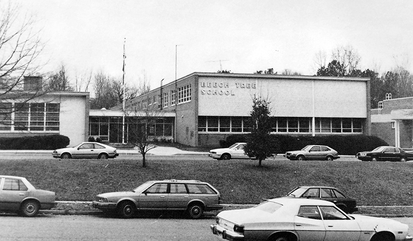 Black and white photograph of the front exterior of Beech Tree Elementary School from our 1986 to 1987 yearbook. The cafeteria and main entrance are visible on the left, the two-story classroom wing at center, and the gymnasium addition is visible on the right. Cars are parked in the bus loop and on the street in front of our building.