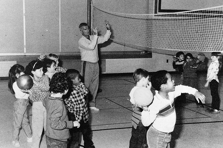 Black and white photograph from our 1986 to 1987 yearbook showing teacher Richard Simonds working with students. A volleyball net has been strung across the gymnasium and two groups of students on either side of the net are tossing balls back and forth across the net to each other.