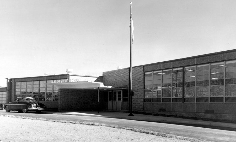 Black and white photograph of the front exterior of Masonville Elementary School. The building is a single-story structure a classroom wing on the right and cafeteria on the left. A 1950s era car is parked in front of the school. 