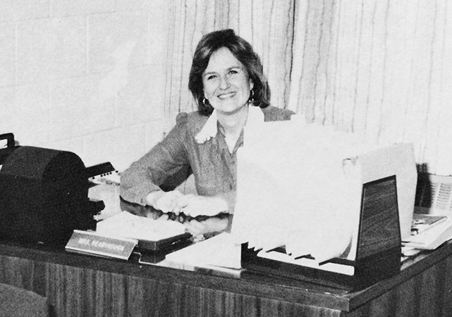 Black and white photograph of Principal Readyhough from our 1984 to 1985 yearbook. She is seated at her desk and is looking up from her work.