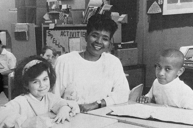 Black and white photograph of Principal Bradley from our 1989 to 1990 yearbook. She is seated at a table speaking with two students about their work.
