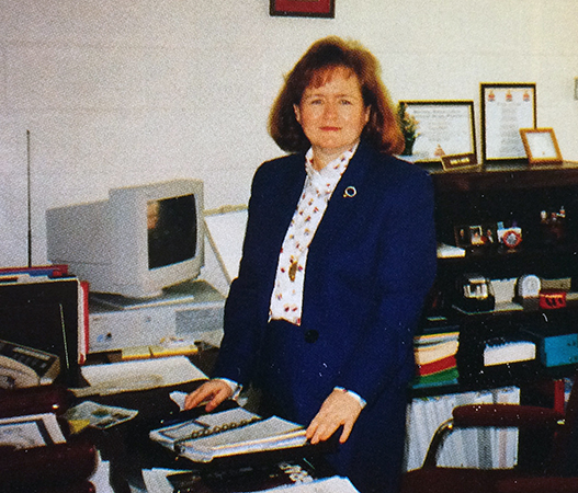Color photograph of Naomi Martin from our 1995 to 1996 yearbook. She is in her office, standing behind her desk. A bookshelf and computer a visible behind her. 