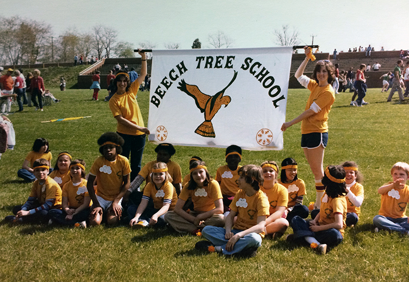 Color photograph of students on the lawn behind Beech Tree Elementary School. The picture was probably taken during the spring time, during one of the annual Fun Fairs. Based on the clothing and hair styles, the time period appears to be the mid or late 1970s. The students are all wearing yellow shirts. Two teachers are holding up a large white banner imprinted with the words Beech Tree School in black, and with an illustration of a bird in flight in the center. The bird is painted yellow and black. Perhaps this was an early mascot for our school before the beaver.