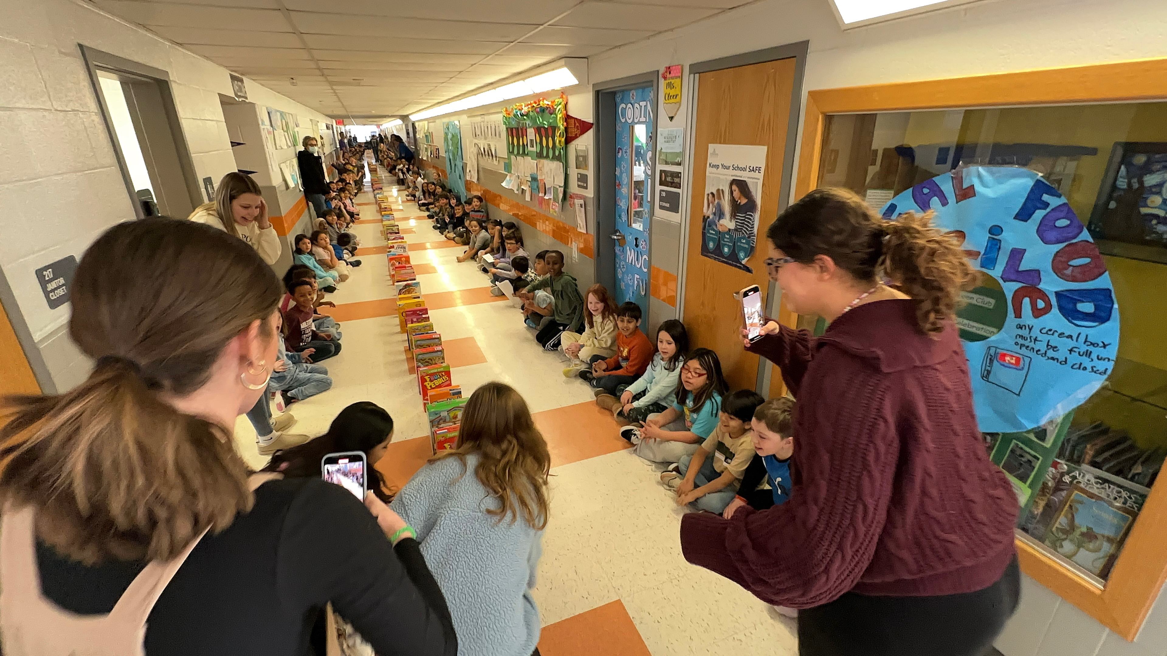 Getting ready the start the cereal domino.  Students sitting against the wall in hallway while a long line of cereal is line up like dominos.